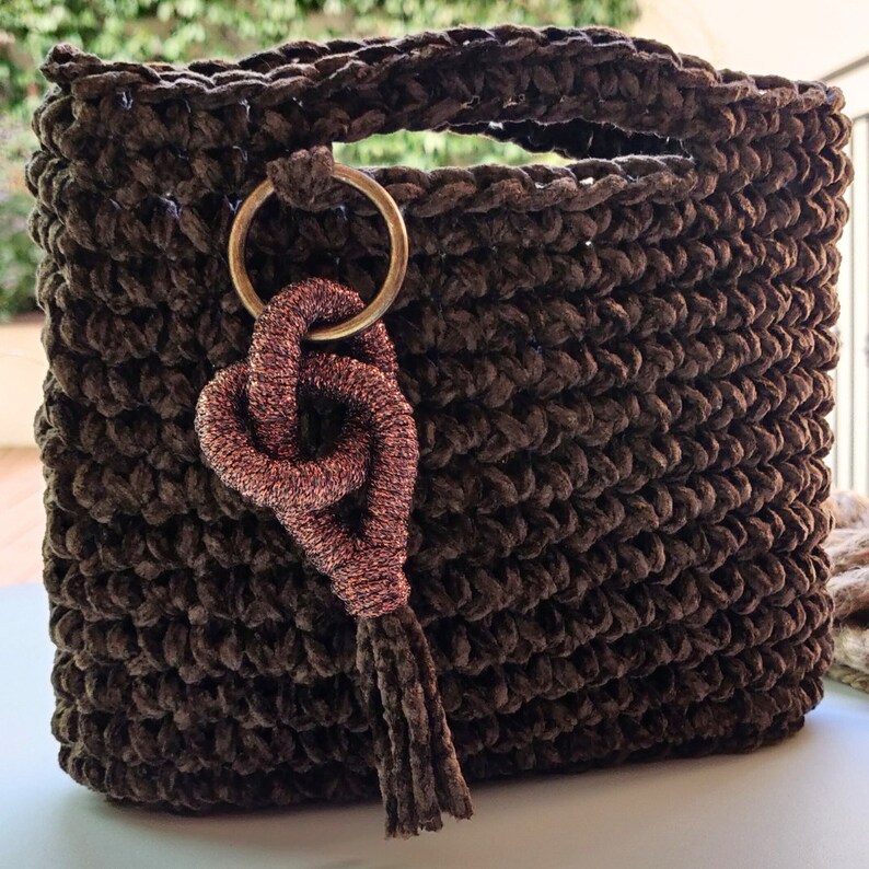 May include: A brown crocheted bag with a gold ring and a pink tassel. The bag is made of a soft, fuzzy material.