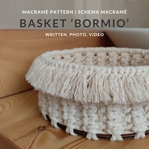 May include: A white macrame basket with a fringe trim. The basket is made with natural cotton cord and has a wooden base. The basket is sitting on a wooden surface.