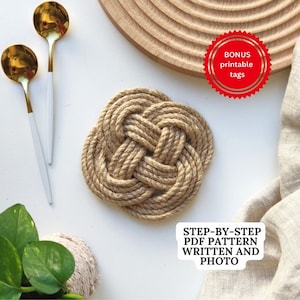 May include: A close-up of a brown rope coaster with a knot design. The coaster is on a white surface with a wooden cutting board and a white linen cloth in the background. The image also includes two gold spoons with white handles and a small potted plant with green leaves. The text "BONUS printable tags" is in a red circle on the top right of the image. The text "STEP-BY-STEP PDF PATTERN WRITTEN AND PHOTO" is in a white rectangle on the bottom right of the image.