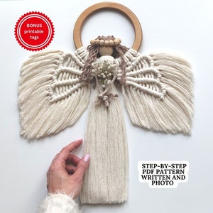 May include: A macrame angel ornament with a wooden ring, white and brown yarn, and a small bouquet of white flowers. The angel has a wooden head with brown yarn hair and a wooden bead crown. The ornament is hanging against a white background.