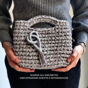 May include: A crocheted handbag in a light gray color with a silver tassel. The bag has a crocheted handle and a silver charm.