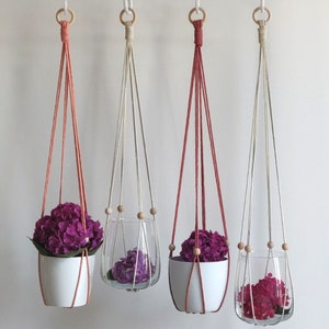 May include: Three macrame plant hangers with wooden beads. The hangers are made with natural colored cord and are hanging from the ceiling. Each hanger has a different colored pot, one is white, one is red, and one is clear glass. The pots are filled with purple flowers.
