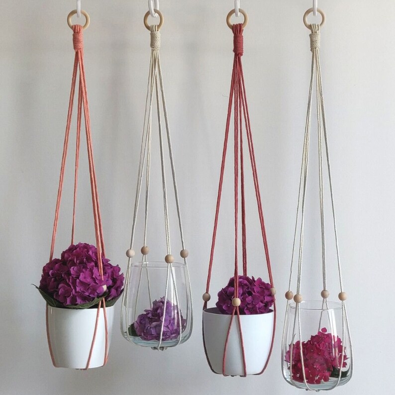 May include: Three macrame plant hangers with wooden beads. The hangers are made with natural colored cord and are hanging from the ceiling. Each hanger has a different colored pot, one is white, one is red, and one is clear glass. The pots are filled with purple flowers.