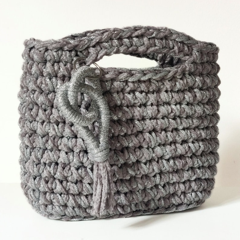 May include: A gray crocheted tote bag with a silver glitter finish and a silver tassel. The bag has a single handle and a silver key chain.