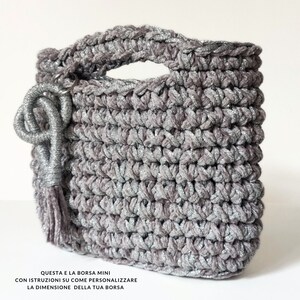 May include: A small, gray crocheted bag with silver glitter. The bag has a top handle and a tassel charm. The text on the bag reads: "QUESTA E LA BORSA MINI CON ISTRUZIONI SU COME PERSONALIZZARE LA DIMENSIONE DELLA TUA BORSA"