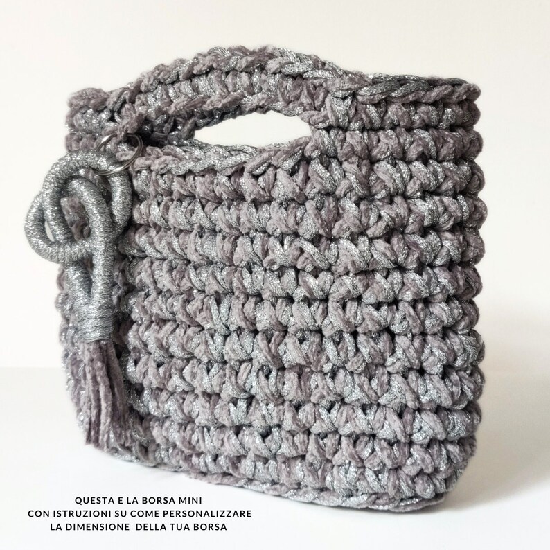 May include: A small, gray crocheted bag with silver glitter. The bag has a top handle and a tassel charm. The text on the bag reads: "QUESTA E LA BORSA MINI CON ISTRUZIONI SU COME PERSONALIZZARE LA DIMENSIONE DELLA TUA BORSA"