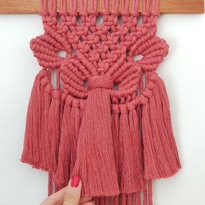 Pode incluir: Um pendurado de macramé rosa com um design floral e franjas. O pendurado é feito com cordão de algodão e está preso a uma barra de madeira.