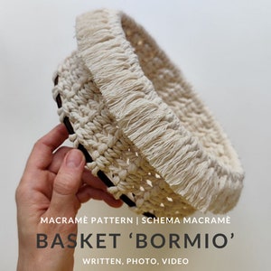 May include: A white macrame basket with a fringe trim. The basket is being held by a hand. The text "MACRAMÉ PATTERN | SCHEMA MACRAMÉ BASKET 'BORMIO' WRITTEN, PHOTO, VIDEO" is at the bottom of the image.
