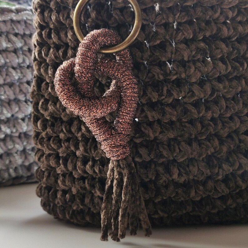 May include: A brown crocheted bag with a copper-colored tassel. The tassel is attached to a ring and has a heart-shaped design.