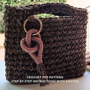 Può includere: Una borsa tote all'uncinetto marrone scuro con un attacco per manico circolare color oro. Un nappa decorativa annodata color rame pende dall'impugnatura. La borsa ha una forma quadrata e un aspetto strutturato. Testo sull'immagine: CROCHET PDF PATTERN, STEP BY STEP INSTRUCTIONS WITH PHOTOS.