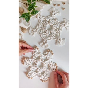 Könnte beinhalten: Zwei weiße Makramee-Schneeflocken mit Holzperlen. Die Schneeflocken hängen an einer weißen Oberfläche.