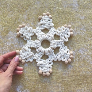 Puede incluir: Un copo de nieve blanco tejido a crochet con cuentas de madera alrededor de los bordes. El copo de nieve está sobre un fondo dorado.