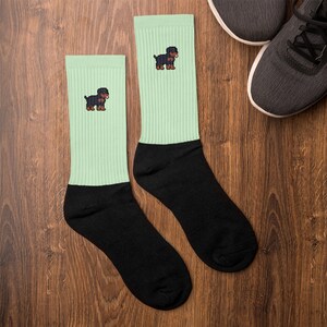 Könnte beinhalten: Ein Paar mintgrüne und schwarze Socken mit einem pixeligen Rottweiler-Hundedesign. Die Socken sind gerippt und haben eine schwarze Spitze und Ferse. Ein Paar graue Turnschuhe sind im Hintergrund auf einer Holzoberfläche zu sehen.