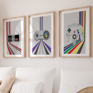 Puede incluir: Tres impresiones enmarcadas de mandos de videojuegos retro. La impresión de la izquierda presenta un mando NES gris y rojo con un fondo gris y rayas de colores. La impresión del medio presenta un mando Super Nintendo gris con un fondo gris y rayas de colores. La impresión de la derecha presenta un mando Nintendo 64 gris con un fondo gris y rayas de colores.