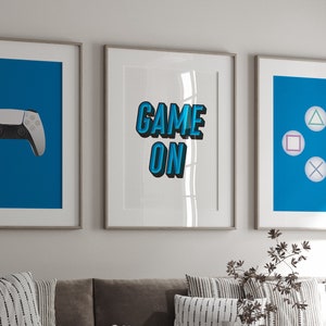 May include: Three framed prints with a blue background. The left print features a white video game controller. The middle print has the text "GAME ON" in blue. The right print has four blue circles with a triangle, square, circle, and cross inside.