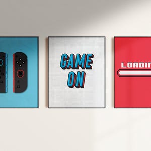 May include: Three framed prints with a gaming theme. The left print features a blue background with two black Nintendo Switch Joy-Con controllers. The middle print has a white background with the text "GAME ON" in blue and red. The right print has a red background with the text "LOADING..." and a white progress bar.