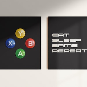 May include: Two black framed prints on a white wall. The print on the left has four colorful buttons with the letters X, Y, A, and B. The print on the right has the text "EAT SLEEP GAME REPEAT" in white.