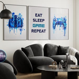 May include: Three framed art prints with a gaming theme. Each print features a blue watercolor design of a game controller or the words "EAT SLEEP GAME REPEAT". The prints are displayed above a dark gray sofa in a modern living room setting.