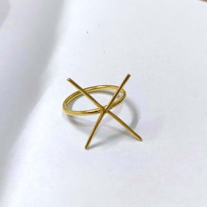 Peut inclure: Une bague dorée avec un sertissage en forme de X. L'anneau est un cercle simple et lisse. Le sertissage comporte quatre griffes s'étendant de l'anneau, conçues pour maintenir une pierre précieuse. La bague est présentée sur une surface blanche.