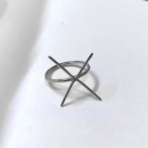 Könnte beinhalten: Ein silberfarbener Ring mit kreuzförmiger Fassung. Der Ring ist dünn und kreisförmig, mit vier Zinken, die nach oben ragen, um einen Edelstein zu halten. Der Ring ist vor einem weißen Hintergrund platziert.