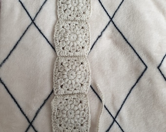 May include: A handmade, off-white crochet bookmark featuring a series of four square granny squares and a pointed end. The bookmark is made of a soft, textured yarn and is lying on a white blanket with a dark blue diamond pattern.