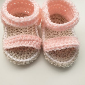 Könnte beinhalten: Ein Paar gehäkelte Babysandalen in Rosa und Beige. Die Sandalen haben einen Riemen über dem Fuß und einen weißen Rand um die Sohle.