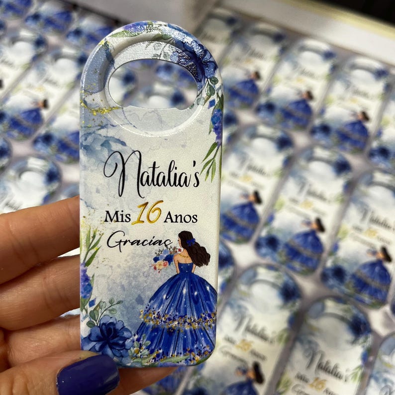 Op de afbeelding: Een witte flessenopener met een blauw en gouden bloemenontwerp. De tekst "Natalia's Mis 16 Anos Gracias" is op de flessenopener gedrukt. Een cartoonafbeelding van een meisje in een blauwe jurk is ook op de flessenopener.