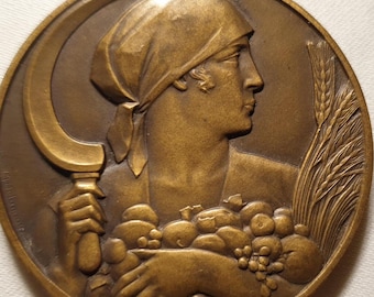 Puede incluir: Escultura en relieve circular de color bronce que representa a una mujer de perfil con pañuelo, sosteniendo una hoz y una cesta de frutas. Espigas de trigo visibles a la derecha. Diseño de estilo vintage.