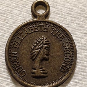 Peut inclure: Un pendentif circulaire de couleur bronze. Il porte l'inscription "QUEEN ELIZABETH THE SECOND" sur le bord et un profil d'une personne portant une couronne de laurier. Une petite boucle est située en haut.