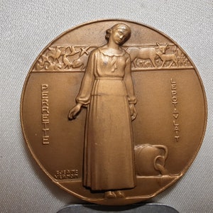 May include: Bronze-colored round medal featuring a woman in a long dress, with the words "PERPETE" and "LEPOTAV LAT" inscribed. The medal also includes images of cows and other figures, with the artist's name, "JEANNE VERNON", at the bottom.