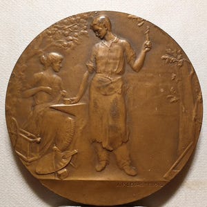 May include: A bronze medal depicting a man and woman working together. The man is holding a hammer and the woman is working on a spinning wheel. The medal is signed "J.P. Legastelois".