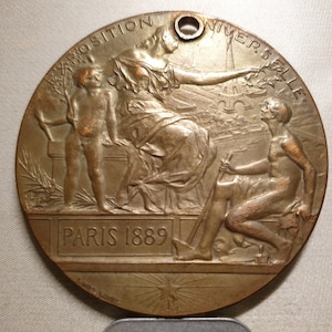 Può includere: Medaglia di bronzo commemorativa dell'Esposizione Universale di Parigi del 1889. La medaglia raffigura una donna che tiene un ramo di ulivo, un giovane ragazzo che tiene un bastone e un uomo che tiene uno strumento. L'iscrizione "Exposition Universelle Paris 1889" è visibile sulla medaglia.