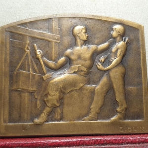 Pode incluir: Escultura retangular de bronze que retrata duas figuras. Uma figura está sentada, segurando um martelo e tenazes perto de uma forja. A segunda figura está em pé, com um braço em volta da figura sentada. A escultura é assinada "E. BLIN".