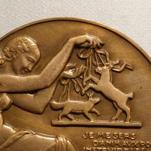 May include: A bronze medal with a relief sculpture of a woman holding a branch with a deer and a fox. The inscription reads "Je me sers d'animaux pour instruire les..."