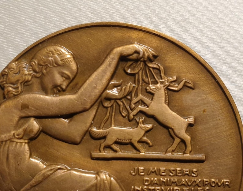 May include: A bronze medal with a relief sculpture of a woman holding a branch with a deer and a fox. The inscription reads "Je me sers d'animaux pour instruire les..."