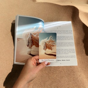 K&ouml;nnte beinhalten: Die Hohepriesterin Tarotkarte mit einem wei&szlig;en und blauen Design. Die Karte zeigt eine Frau in einem wei&szlig;en Gewand, die in einer W&uuml;stenlandschaft steht. Der Kartentext lautet "Die Hohepriesterin" und "Geheimnis - Weisheit - Geheimnisse".