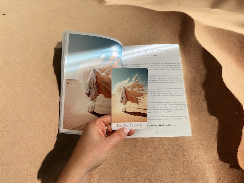 K&ouml;nnte beinhalten: Die Hohepriesterin Tarotkarte mit einem wei&szlig;en und blauen Design. Die Karte zeigt eine Frau in einem wei&szlig;en Gewand, die in einer W&uuml;stenlandschaft steht. Der Kartentext lautet "Die Hohepriesterin" und "Geheimnis - Weisheit - Geheimnisse".