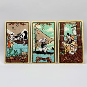 May include: Three tarot cards with a nautical theme. The cards feature illustrations of a devil, a hermit, and a high priestess. Each card has a cream-colored border and the card titles are printed at the bottom.