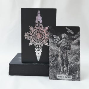 Puede incluir: Una caja de cartas de tarot negra con un diseño de mandala holográfico y una carta de tarot titulada Page of Blades. La carta muestra una ilustración en escala de grises de una persona en un campo con pájaros bajo un cielo oscuro y nublado.