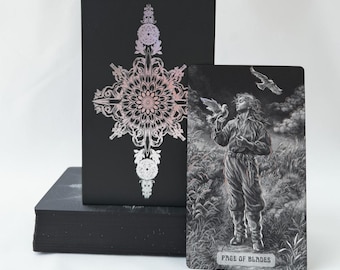 May include: A black tarot card box featuring a holographic mandala design and a single tarot card titled Page of Blades. The card displays a grayscale illustration of a person standing in a field with birds, set against a dark, cloudy sky.