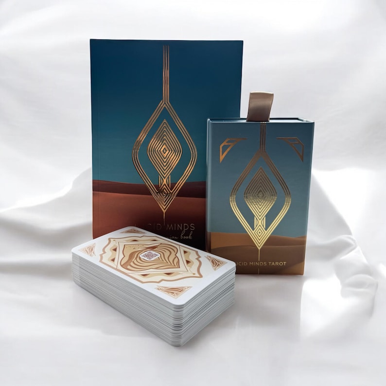 K&ouml;nnte beinhalten: Eine blaugr&uuml;ne Schachtel mit goldenen Akzenten und dem Text "Good Minds Tarot" auf der Vorderseite. Die Schachtel befindet sich neben einem Stapel Tarotkarten mit einem gold-braunen Design. Die Karten liegen auf einer wei&szlig;en Oberfl&auml;che.