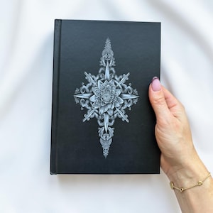 Puede incluir: Un libro de tapa dura negro con una ilustración blanca intrincada centrada en la portada. El diseño muestra una flor de loto detallada rodeada de elementos en forma de espada y patrones de filigrana simétricos.