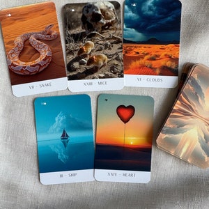 May include: Five tarot cards with colorful images and text. The cards are laid out in a row on a beige fabric surface. The cards are labeled with Roman numerals and the names of the cards: VII - Snake, XXIII - Mice, VI - Clouds, III - Ship, XXIV - Heart.