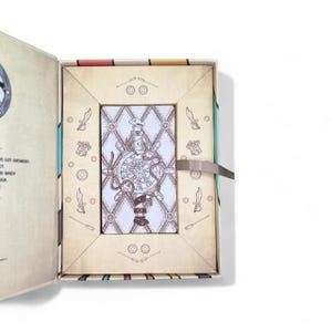 May include: Open tarot card box with a card featuring a nautical design. The card shows a ship's wheel with a lighthouse and the word "PINK". The box is beige with decorative elements and a ribbon closure.