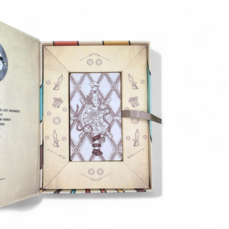 May include: Open tarot card box with a card featuring a nautical design. The card shows a ship's wheel with a lighthouse and the word "PINK". The box is beige with decorative elements and a ribbon closure.