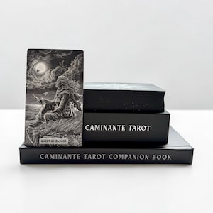 May include: A black and white tarot card titled Queen of Blades featuring a figure sitting on a cliff under a moonlit sky, placed next to a black box labelled Caminante Tarot and a black book titled Caminante Tarot Companion Book.