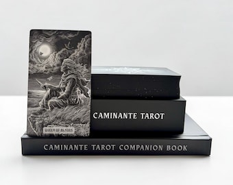 May include: A black and white tarot card titled Queen of Blades featuring a figure sitting on a cliff under a moonlit sky, placed next to a black box labeled Caminante Tarot and a black book titled Caminante Tarot Companion Book.