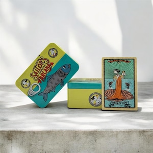Puede incluir: Una lata amarilla y verde azulado de "Sailor's Arcana" con una ilustración de pez y el texto "Sailor's Arcana". Una carta del tarot con el texto "Queen of Cups" está al lado de la lata. También hay una segunda lata amarilla y verde azulado.