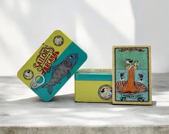 Könnte beinhalten: Eine gelb-türkisfarbene "Sailor's Arcana"-Dose mit einer Fischillustration und dem Text "Sailor's Arcana". Eine Tarotkarte mit dem Text "Queen of Cups" befindet sich neben der Dose. Eine zweite gelb-türkisfarbene Dose ist ebenfalls vorhanden.