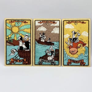 May include: Three tarot cards featuring cartoon characters. "The Sun" card shows a sunny beach scene. "Three of Swords" depicts a boat in the rain. "Strength" shows a character riding a fish. Each card has a decorative border and banner.